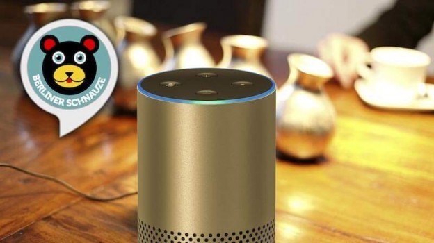 Ein Smart Speaker mit dem Alexa Skill „Berliner Schnauze“ steht zwischen mehreren silbernen Objekten auf einem Tisch, während sich im Hintergrund Menschen unterhalten.